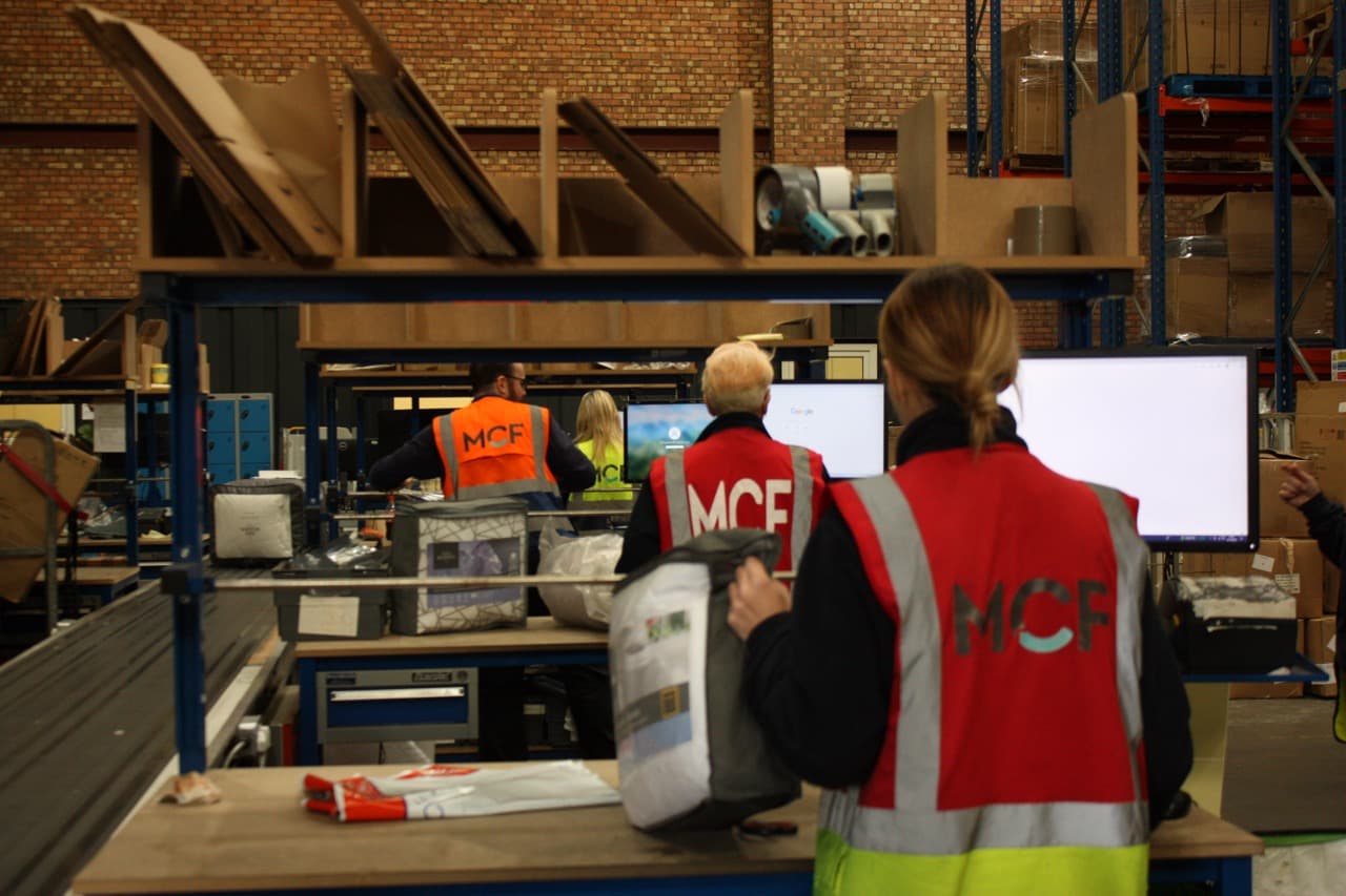 MCF warehouse team members in branded high-visibility vests working at packing stations inside the MCF fulfilment centre in Liverpool.
