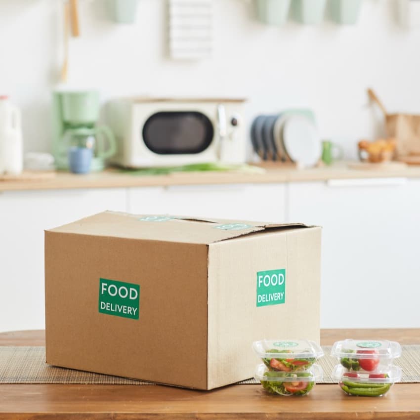 Background image of cardboard box with food standing on wooden table in white kitchen interior, food delivery service, copy space