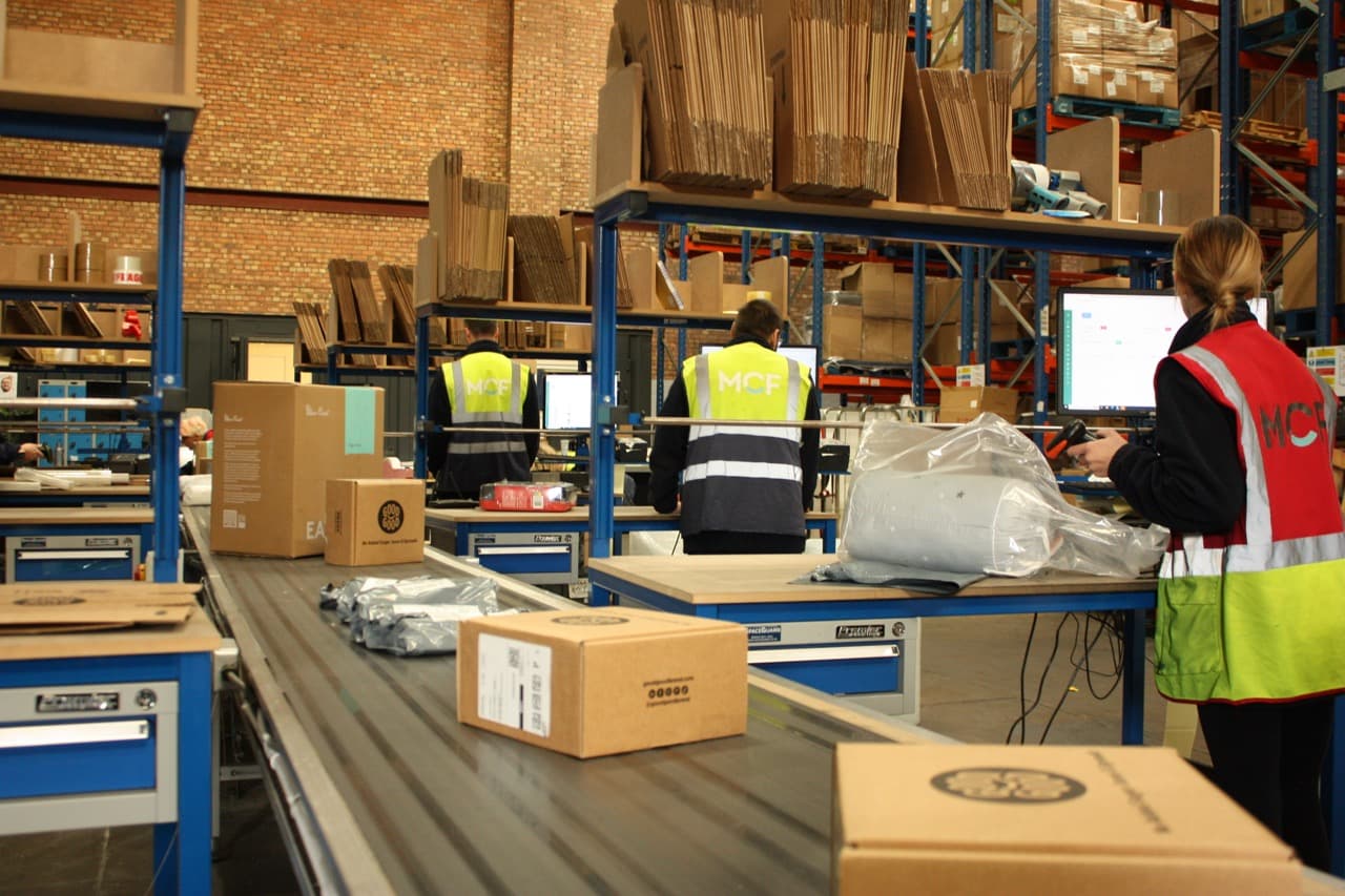 Fulfilment team processing and scanning orders at packing stations along a conveyor belt in a busy warehouse.